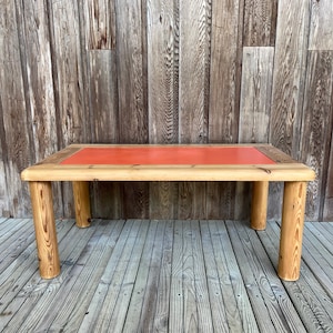 Peut inclure: Une table basse rectangulaire en bois avec un panneau central orange vif. La table a quatre pieds cylindriques épais et une finition en bois naturel. La table mesure environ 122 cm de long et 61 cm de large.