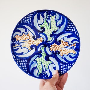 Puede incluir: Un plato redondo de cerámica pintado a mano con un diseño vibrante. El plato presenta un patrón azul y blanco con un diseño central de puntos marrones y verdes. El borde tiene un patrón de cuadros azules y blancos. El plato está sostenido por una mano.