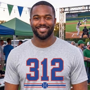 May include: A light grey short-sleeved t-shirt featuring the number 312 in large blue font with orange outlines, positioned above a horizontal graphic of two blue lines with a small blue baseball icon centered between them on the chest.