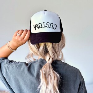 Könnte beinhalten: Eine schwarz-weiße Trucker-Kappe mit dem Wort "CUSTOM" in Schwarz auf der Vorderseite. Die Kappe wird von einer Person mit langen blonden Haaren getragen.