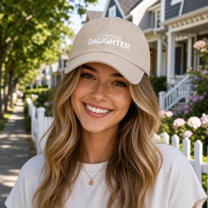May include: A beige baseball cap with the words "Favorite Daughter" embroidered in white script. The cap is worn by a person with long blonde hair, smiling in front of a white picket fence and houses.