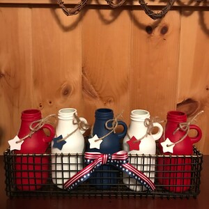 May include: Five painted glass milk bottles in a wire basket. The bottles are red, white, and blue with a star on each. The basket is decorated with a red, white, and blue ribbon.