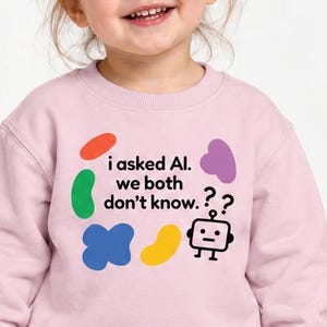 May include: A light pink crewneck sweatshirt featuring a graphic print with the text "i asked AI. we both don't know." accompanied by a small black robot icon, two question marks, and various colorful abstract shapes in red, green, blue, yellow, and purple.