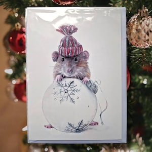 May include: A Christmas card featuring a detailed illustration of a brown rat wearing a red and white striped knit hat. The rat is holding a large white ornament with snowflake designs. The card is set against a blurred Christmas tree background.