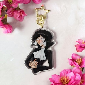 Peut inclure: Un porte-clés en acrylique transparent avec une illustration de style anime d'une personne aux cheveux bouclés noirs, une cravate blanche et un costume noir. Le porte-clés a un fermoir en forme d'étoile dorée et un anneau doré. Des fleurs roses sont en arrière-plan.
