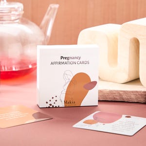 May include: A white box with the text "Pregnancy Affirmation Cards" and a line drawing of a pregnant woman. The box is on a pink surface with a few cards from the deck visible.
