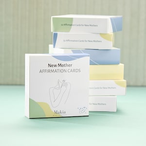 May include: A white box with the text "New Mother Affirmation Cards" and a line drawing of a person. The box is on a light blue surface. The text "Makia Mind Mantras" is also on the box.