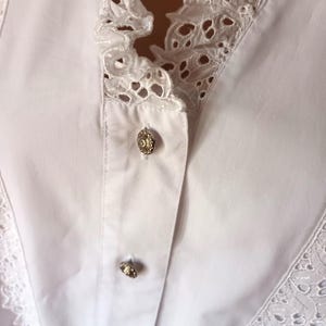 Puede incluir: Primer plano de una blusa blanca con un ribete de encaje calado decorativo a lo largo del cuello y la parte delantera. La blusa presenta tres botones dorados ornamentados en la parte delantera. La tela parece ser un algod&oacute;n crujiente y ligero.