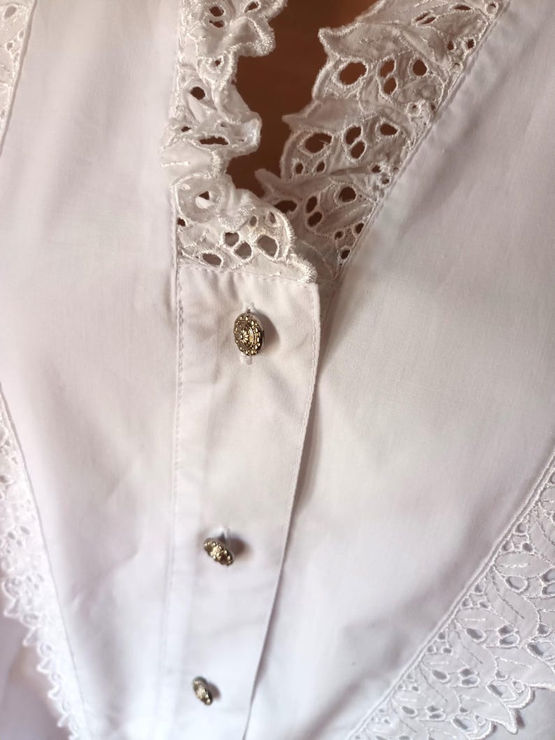 Puede incluir: Primer plano de una blusa blanca con un ribete de encaje calado decorativo a lo largo del cuello y la parte delantera. La blusa presenta tres botones dorados ornamentados en la parte delantera. La tela parece ser un algod&oacute;n crujiente y ligero.