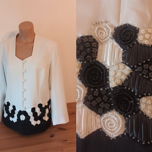 May include: A white blazer with a black and white geometric pattern on the bottom hem. The blazer has a V-neck and a button closure.