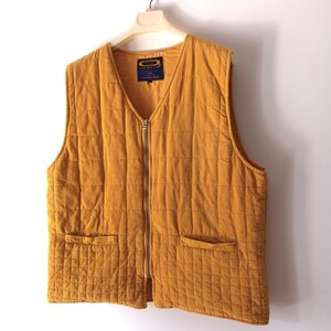 May include: Mustard-coloured quilted vest with a V-neck and a full-length zip. The vest has two front pockets and is hanging on a hanger. The fabric has a corduroy texture. A small label is visible near the neckline.