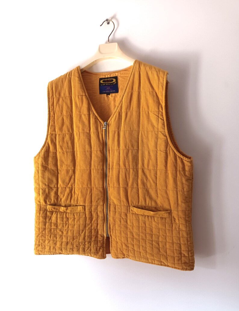 May include: Mustard-coloured quilted vest with a V-neck and a full-length zip. The vest has two front pockets and is hanging on a hanger. The fabric has a corduroy texture. A small label is visible near the neckline.