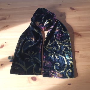 Puede incluir: Bufanda de terciopelo negro con un estampado floral en dorado, morado y rosa. La bufanda está drapeada sobre una superficie de madera, mostrando su textura suave y su intrincado diseño. La bufanda tiene una etiqueta en el extremo.