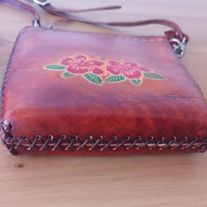 May include: A brown leather crossbody bag with a floral design. The bag has a stitched edge and a small metal clasp. The bag is signed "Gino".