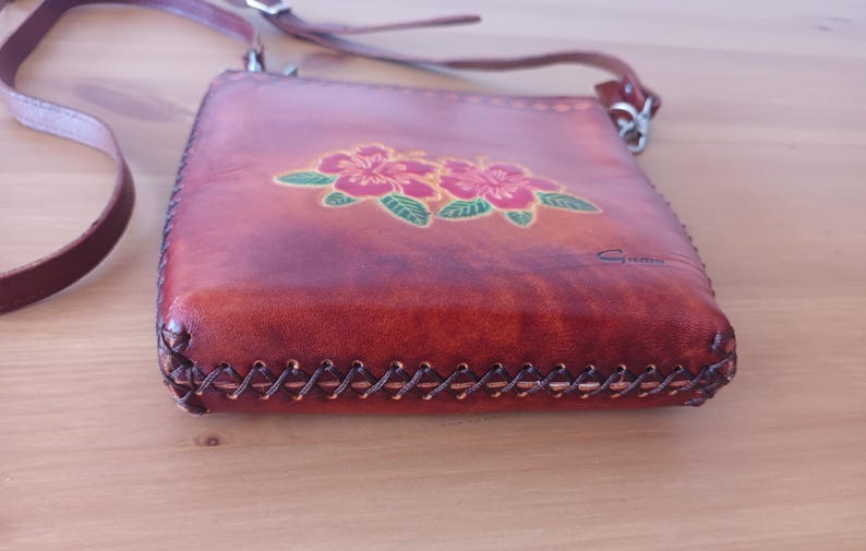 May include: A brown leather crossbody bag with a floral design. The bag has a stitched edge and a small metal clasp. The bag is signed "Gino".