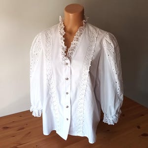 Puede incluir: Una blusa blanca con cuello alto con volantes y mangas largas. La blusa presenta intrincados detalles de encaje en la parte delantera, las mangas y el cuello. Las mangas tienen un ligero abullonado en los hombros y est&aacute;n recogidas en los pu&ntilde;os.