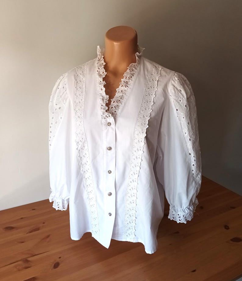 Puede incluir: Una blusa blanca con cuello alto con volantes y mangas largas. La blusa presenta intrincados detalles de encaje en la parte delantera, las mangas y el cuello. Las mangas tienen un ligero abullonado en los hombros y est&aacute;n recogidas en los pu&ntilde;os.