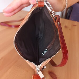 May include: A small, light brown leather purse with a zipper closure and a long strap. The purse is open and the inside is lined with a dark brown fabric.