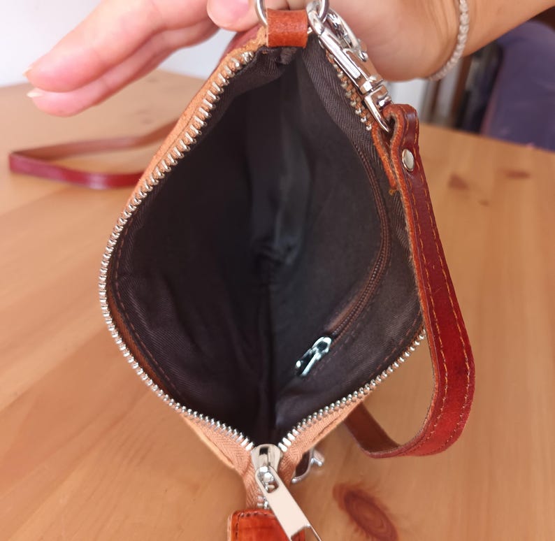 May include: A small, light brown leather purse with a zipper closure and a long strap. The purse is open and the inside is lined with a dark brown fabric.