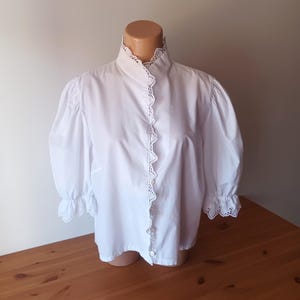 Pode incluir: Uma blusa branca de bot&otilde;es com gola alta, mangas bufantes volumosas e um delicado acabamento em renda festonada com ilh&oacute;s ao longo da carcela frontal, gola e punhos. A pe&ccedil;a est&aacute; exibida num torso de manequim.