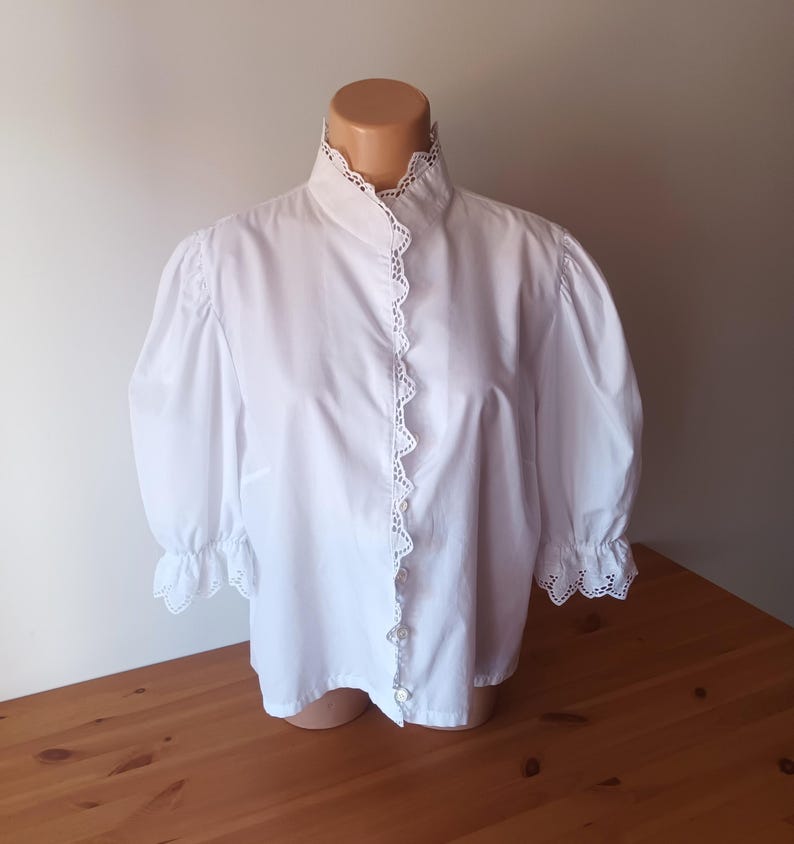Pode incluir: Uma blusa branca de bot&otilde;es com gola alta, mangas bufantes volumosas e um delicado acabamento em renda festonada com ilh&oacute;s ao longo da carcela frontal, gola e punhos. A pe&ccedil;a est&aacute; exibida num torso de manequim.