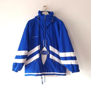 Könnte beinhalten: Blaue und weiße Vintage-Anorakjacke mit Kapuze und halbem Reißverschluss. Die Jacke hat weiße Streifen an den Ärmeln und ein weißes Design auf der Vorderseite. Das Wort "BOGNER" ist auf der Brust sichtbar.