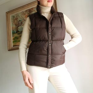 Puede incluir: Un chaleco acolchado marrón con cuello alto y cierre de botones. El chaleco se lleva sobre un jersey de cuello alto color crema y pantalones blancos. El chaleco tiene un diseño acolchado y un bolsillo lateral.