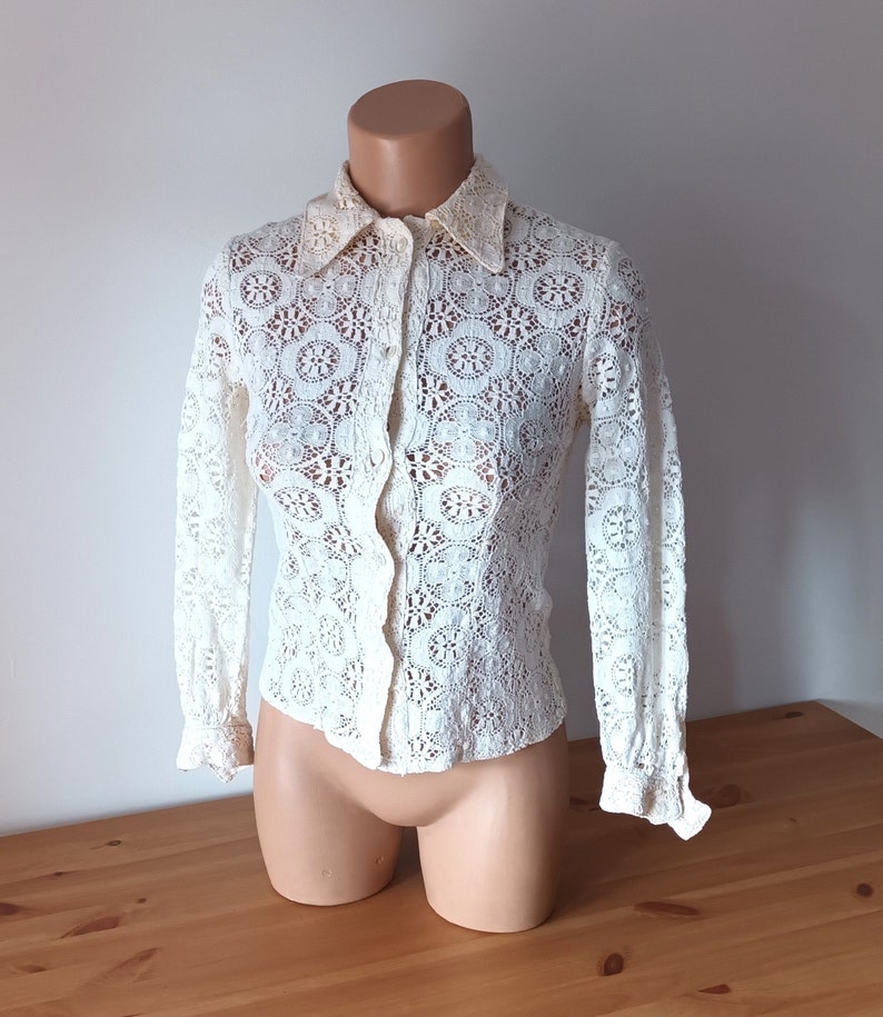 May include: A white crochet blouse with long sleeves and a button-up front. The blouse has a delicate floral pattern and a classic collar.