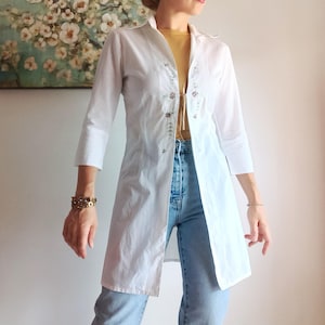 May include: A white long-line button-down shirt featuring three-quarter length sleeves and delicate floral embroidery along the front placket. The shirt is worn open over a mustard yellow top and light blue denim jeans, creating a layered look.