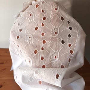 Puede incluir: Tela calada blanca con motivos florales y de puntos. La tela forma parte de una prenda, posiblemente una blusa o un vestido, con un dise&ntilde;o calado detallado. La prenda tiene un cuello con volantes y est&aacute; drapeada sobre una superficie de madera.