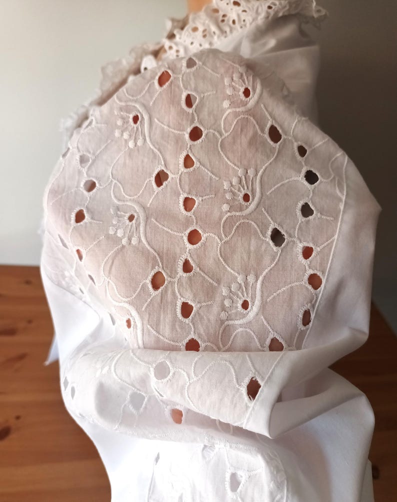 Puede incluir: Tela calada blanca con motivos florales y de puntos. La tela forma parte de una prenda, posiblemente una blusa o un vestido, con un dise&ntilde;o calado detallado. La prenda tiene un cuello con volantes y est&aacute; drapeada sobre una superficie de madera.