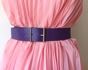 May include: A wide, purple belt with a silver-colored square buckle. The belt has a textured, embossed pattern and is worn over a pink, pleated dress. The belt is a bold fashion accessory.