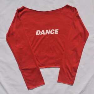 May include: Red long-sleeved shirt with a boat neck and the word "DANCE" printed in white on the front.