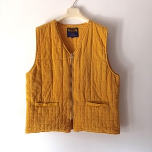 May include: Mustard yellow quilted vest with a V-neck and a full-length zip. The vest has two front pockets and a small label on the inside. The vest is hanging on a white hanger against a white background.