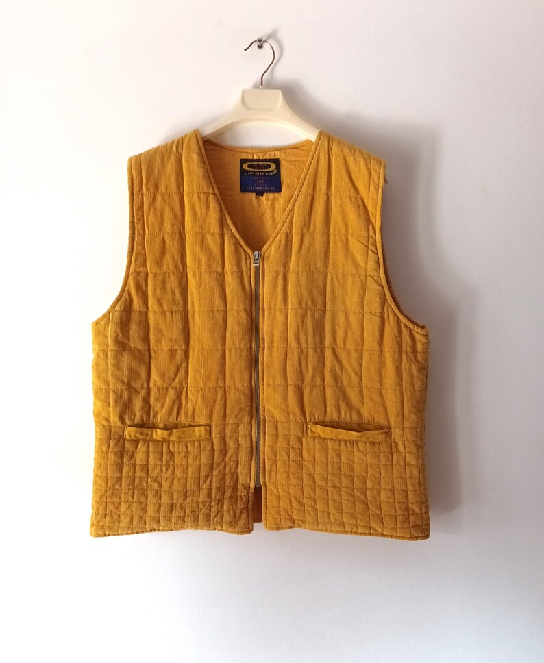 May include: Mustard yellow quilted vest with a V-neck and a full-length zip. The vest has two front pockets and a small label on the inside. The vest is hanging on a white hanger against a white background.