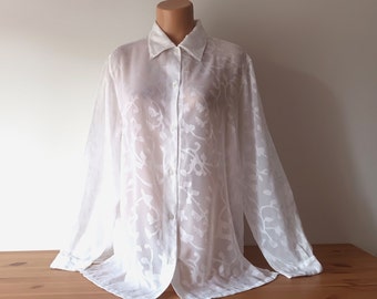 May include: A white, long-sleeved button-down shirt with a floral pattern. The shirt has a classic collar and is displayed on a mannequin. The fabric appears sheer, with the floral design adding texture and visual interest to the garment.