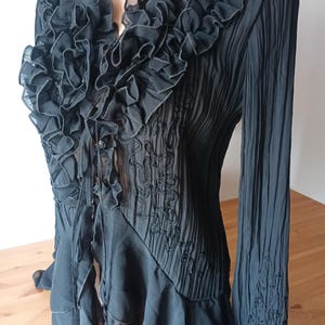 Puede incluir: Blusa negra con cuello con volantes y mangas largas. La blusa presenta un dise&ntilde;o plisado y detalles bordados en el corpi&ntilde;o y las mangas. Los volantes caen en cascada por la parte delantera.