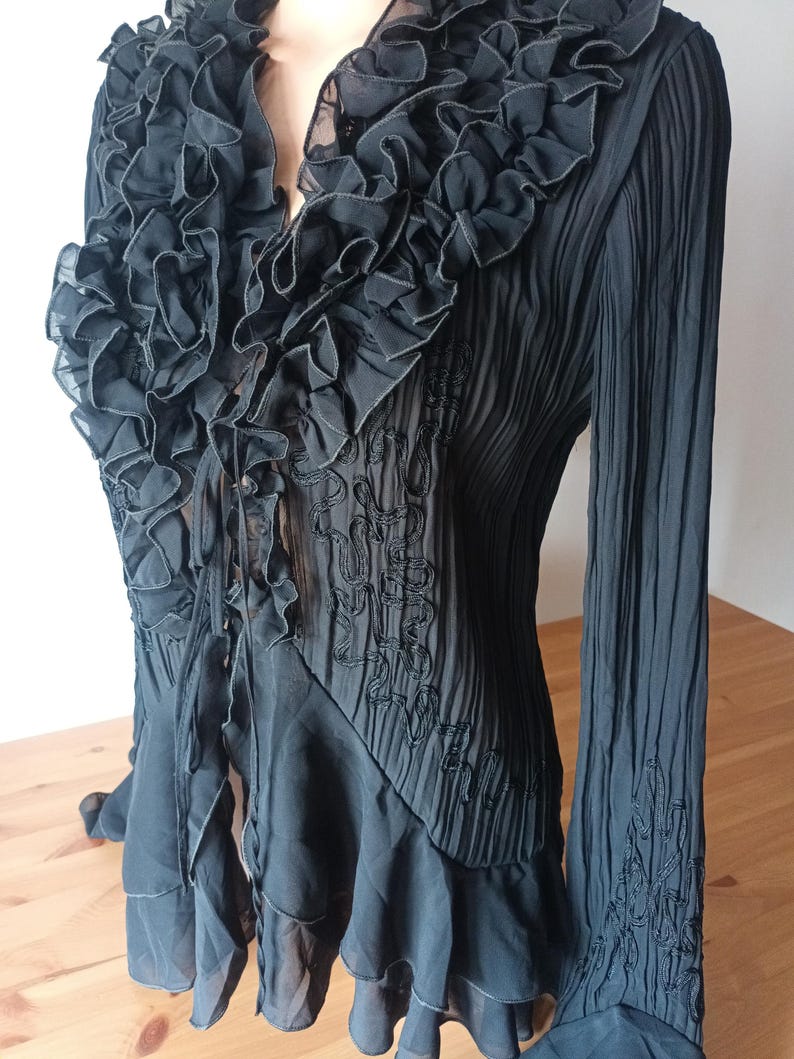 Puede incluir: Blusa negra con cuello con volantes y mangas largas. La blusa presenta un dise&ntilde;o plisado y detalles bordados en el corpi&ntilde;o y las mangas. Los volantes caen en cascada por la parte delantera.