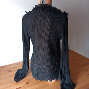 Puede incluir: Blusa negra plisada de manga larga con cuello y pu&ntilde;os con volantes. La blusa se muestra en un maniqu&iacute;, mostrando su tejido texturizado y su dise&ntilde;o. Las mangas tienen un detalle acampanado y con volantes en la mu&ntilde;eca.