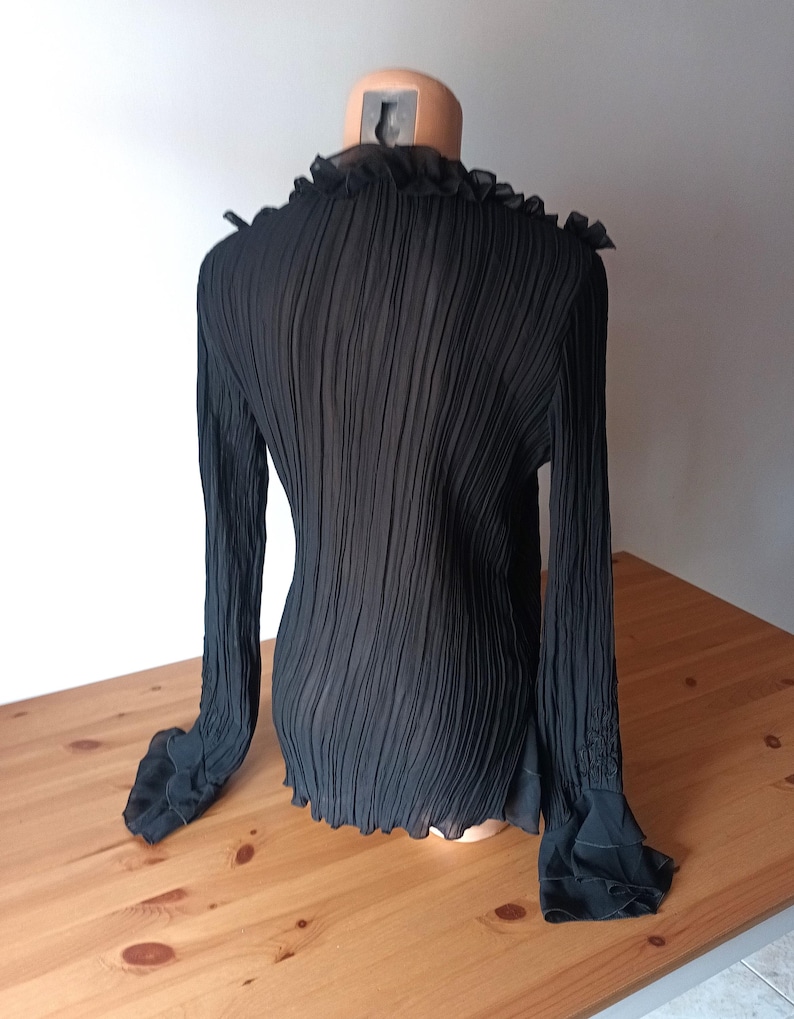 Puede incluir: Blusa negra plisada de manga larga con cuello y pu&ntilde;os con volantes. La blusa se muestra en un maniqu&iacute;, mostrando su tejido texturizado y su dise&ntilde;o. Las mangas tienen un detalle acampanado y con volantes en la mu&ntilde;eca.