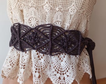 Puede incluir: Un cinturón de macramé marrón oscuro con intrincados patrones tejidos. El cinturón se usa sobre una blusa de encaje color crema, creando un estilo bohemio. El cinturón tiene un cierre de lazo y es ancho.