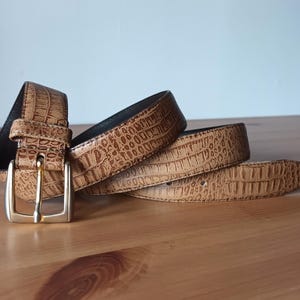 Könnte beinhalten: Ein brauner Gürtel mit einem strukturierten, krokodilartigen Muster. Der Gürtel hat eine goldfarbene Schnalle und liegt auf einer Holzoberfläche. Ein stilvolles Accessoire.