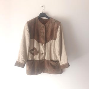 May include: A brown and beige patchwork jacket with a button closure. The jacket has a suede-like texture and features a decorative design on the front.