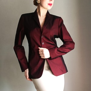 Könnte beinhalten: Ein burgunderfarbener Blazer mit Zwei-Knopf-Verschluss. Der Blazer hat einen subtilen Glanz und eine taillierte Passform. Die Ärmel sind lang und die Revers sind gekerbt. Ein klassisches Oberbekleidungsstück.