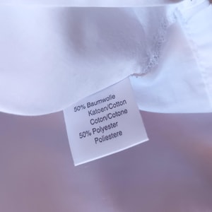 Pode incluir: Uma etiqueta de tecido branco para vestu&aacute;rio com texto impresso em preto que diz 50% Baumwolle, Katoen/Cotton, Coton/Cotone e 50% Polyester, Poliestere. A etiqueta est&aacute; fixada numa superf&iacute;cie t&ecirc;xtil branca lisa com costuras vis&iacute;veis.