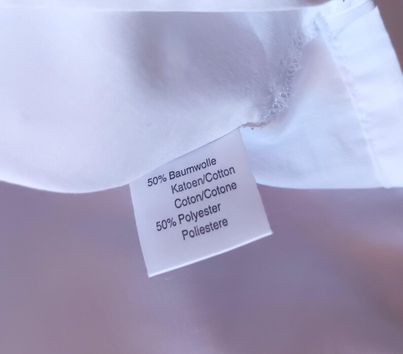 Pode incluir: Uma etiqueta de tecido branco para vestu&aacute;rio com texto impresso em preto que diz 50% Baumwolle, Katoen/Cotton, Coton/Cotone e 50% Polyester, Poliestere. A etiqueta est&aacute; fixada numa superf&iacute;cie t&ecirc;xtil branca lisa com costuras vis&iacute;veis.