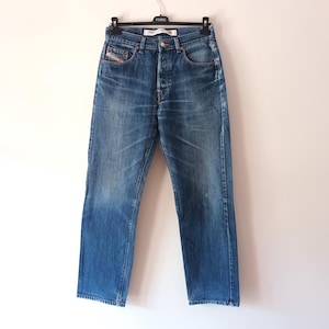 Könnte beinhalten: Eine blaue Jeans aus Denim mit verwaschener Optik, ausgestattet mit einem klassischen Fünf-Taschen-Design, einer Knopfleiste und kupferfarbenen Nieten. Die Jeans hängt an einem schwarzen Kunststoffbügel und zeigt einen geraden Schnitt mit Sitzfalten.