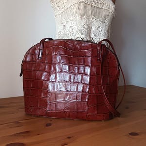 May include: A brown leather handbag with a crocodile-embossed pattern. The bag has a top zipper closure and a long shoulder strap.