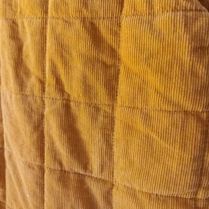 May include: Close-up of a mustard yellow corduroy fabric with a quilted pattern. The fabric has vertical ridges and is divided into square sections by horizontal and vertical stitching. The colour is a warm, earthy tone.