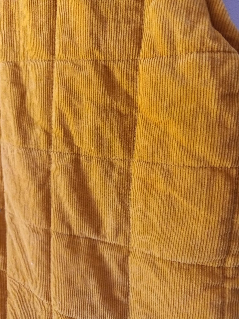 May include: Close-up of a mustard yellow corduroy fabric with a quilted pattern. The fabric has vertical ridges and is divided into square sections by horizontal and vertical stitching. The colour is a warm, earthy tone.